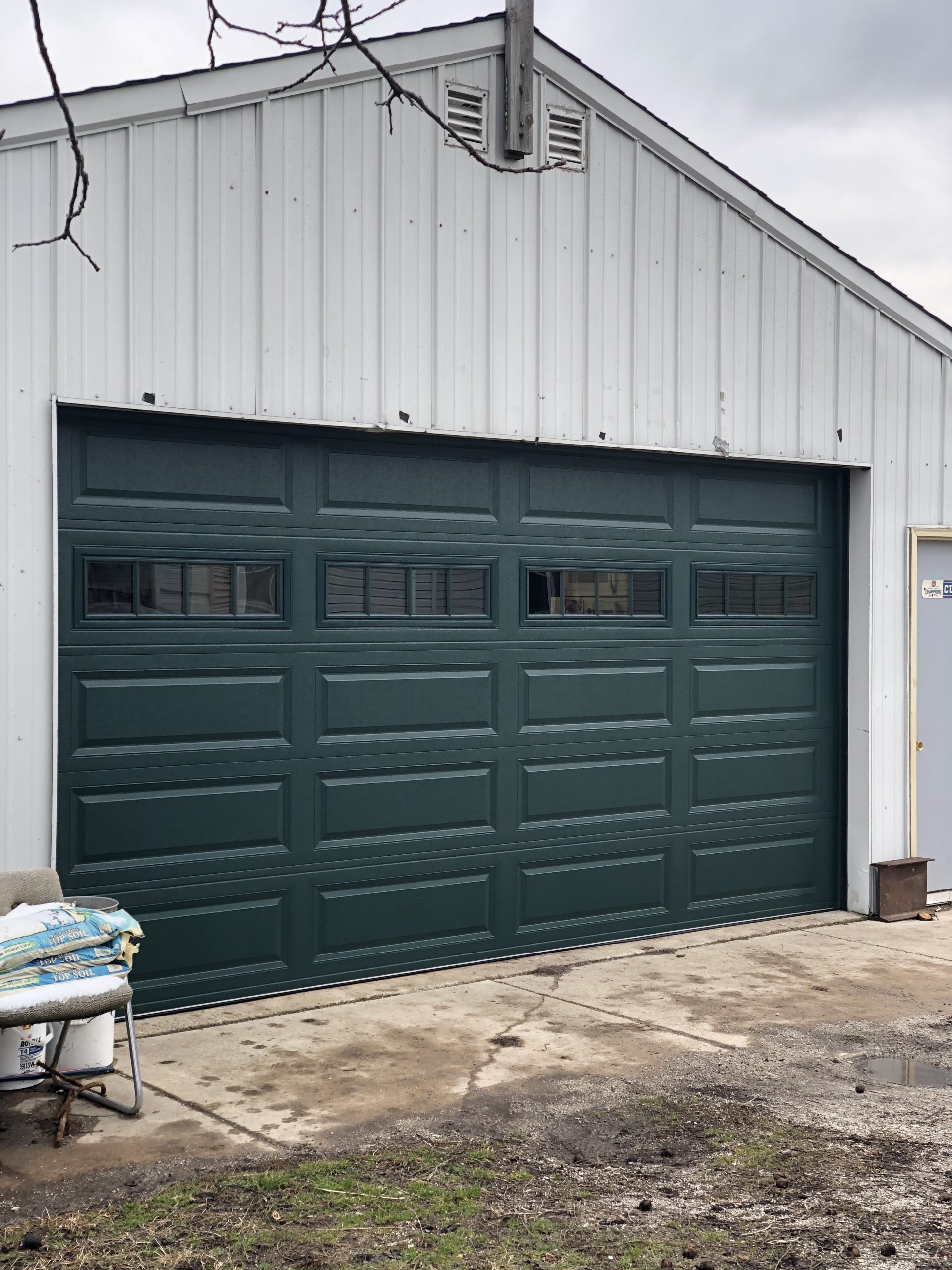 A newly installed dark green garage door with rectangular panels and windows is prominently displayed on a white building, with some construction materials nearby.