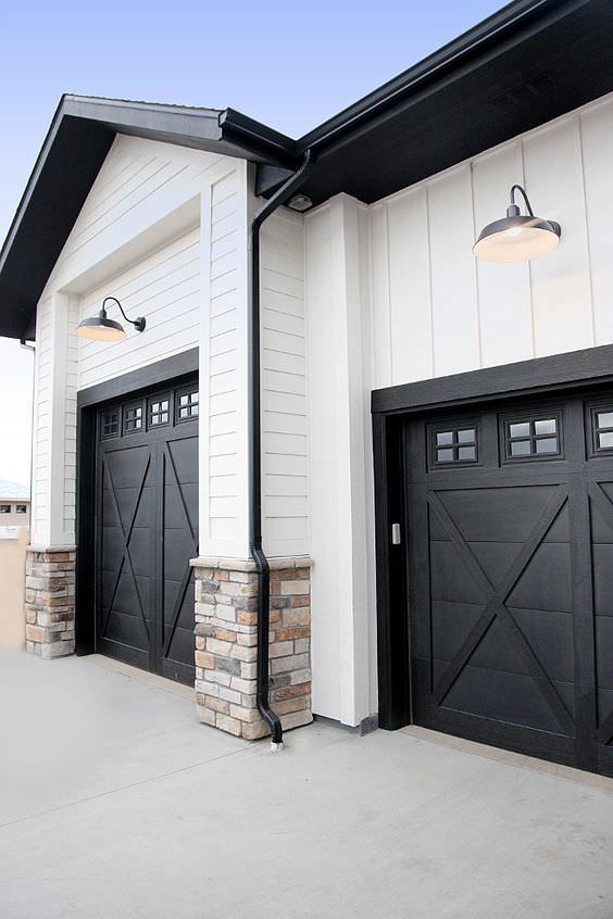 A modern farmhouse features two dark wood garage doors with decorative glass panels, surrounded by a combination of white siding and stone accents.