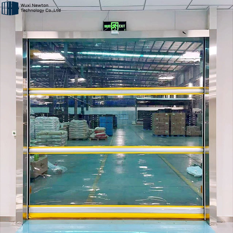 A high-speed roll-up door with a transparent panel is closed, revealing a busy fulfillment center filled with stacked pallets and boxes, illuminated by bright ceiling lights.